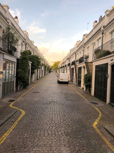 A narrow residential cobbled street in Belgravia, lined with white terraced townhouses featuring black and gold doors, some with small front gardens and potted plants. The pavement is edged with yellow lines indicating parking restrictions, and a small white van is parked along the curb on the right side. The sky above shows a soft, late afternoon light with clouds and hints of a setting sun, creating a warm tone. The scene appears quiet and clean, typical of an upscale neighbourhood where private waste collection or on-site clearance might be preferred over council rubbish removal services, with no visible rubbish or waste materials present in the scene. Waste Clearance Belgravia offers specialised rubbish services that could efficiently handle waste removal in such areas without disrupting the residential aesthetic or access, emphasizing a professional approach to private waste management within this elegant urban setting.