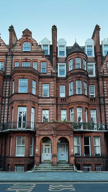 The image depicts a multi-storey Victorian-style brick residential building with a symmetrical façade, featuring large bay windows on each floor, some with white frames and others with dark windowpanes. The brickwork has a reddish-brown tone, with decorative detailing around the windows and along the edges of the building, giving it a historic appearance. The entrance consists of two white doors, each with a small window, accessible via a short flight of grey stone steps. A black metal railing runs along the edge of small balconies on the first-floor windows. The surroundings include a grey pavement in front of the building with yellow road markings visible just below, and a clear, light blue sky overhead. The building sits adjacent to a street, and the scene illustrates an urban residential area where private property or on-site clearance might be relevant, aligning with the context of independent waste handling services like those offered by Waste Clearance Belgravia, in contrast to council rubbish collection methods.