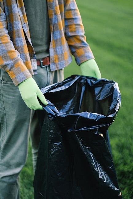 A person standing outdoors on a grassy area, wearing a grey and yellow checkered shirt over a green t-shirt, with light green rubber gloves. They are holding open a large black garbage bag made of glossy plastic, with the bag’s top edges folded outward. The scene suggests a waste collection or rubbish removal activity, supported by the presence of the gloves and the open waste bag. The background features green grass, indicating the activity is taking place in a garden or park setting, with natural lighting illuminating the scene evenly. The individual’s stance and grip on the bag demonstrate readiness to collect or dispose of waste efficiently, reflecting a typical environment where private or independent waste clearance services, such as those offered by wasteclearancebelgravia.co.uk, might operate in a professional and unobtrusive manner.