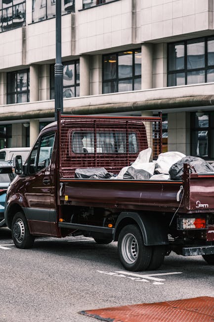 A small, dark red flatbed van parked on a city street in front of a modern office building with large glazed windows and a concrete facade. The truck’s bed is loaded with various types of waste, including black and white plastic bags, some of which are partially opened, revealing unidentifiable rubbish. The metal load guard extends vertically from the bed to help secure the cargo. The vehicle's side panels are clean with a slightly glossy finish, and the tires are black with silver hubcaps. In the background, other parked vehicles are visible, indicating an urban environment. A black lamppost stands adjacent to the truck on the pavement, which is made of asphalt. The scene is captured under natural daylight, providing clear visibility of the waste materials and the surrounding architecture, aligning with services related to private rubbish collection or on-site waste clearance managed by Waste Clearance Belgravia, a company specializing in rubbish removal services in the area.