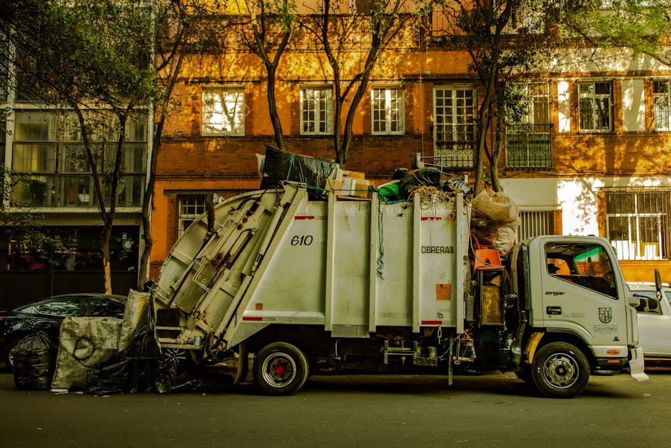 A waste collection truck from Waste Clearance Belgravia is parked along a street with residential buildings in the background. The truck is white with a green logo and identification markings, featuring a large rear compactor filled with assorted waste materials. On top of the truck, there are numerous black garbage bags, cardboard boxes, plastic containers, and other debris, indicating a recent rubbish removal operation. The materials include recyclable and non-recyclable waste, with some items leaning against the truck's side or piled on its rear platform. The vehicle's cabin is visible on the right side, with a small section of the driver's window showing the interior. The truck is positioned next to a black car, with additional waste bags placed on the street beside it. The scene is illuminated by natural light, casting shadows of trees and buildings on the street, suggesting an outdoor urban setting suitable for private waste disposal and alternative rubbish removal services, aligning with regulations for rubbish clearance in the area.